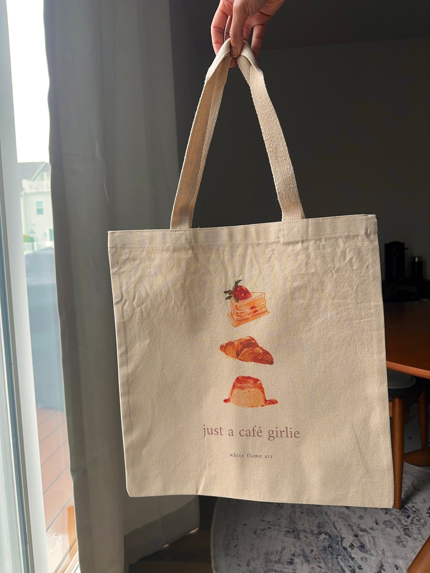 Tote bag with illustrations of pastries and text held by a hand indoors.