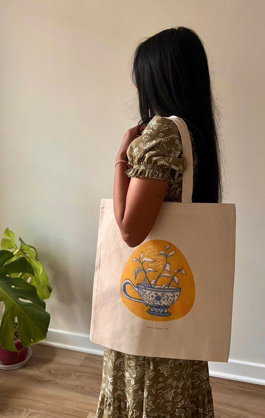 Person holding a tote bag with a design of a teacup and saucer, standing indoors next to a plant.
