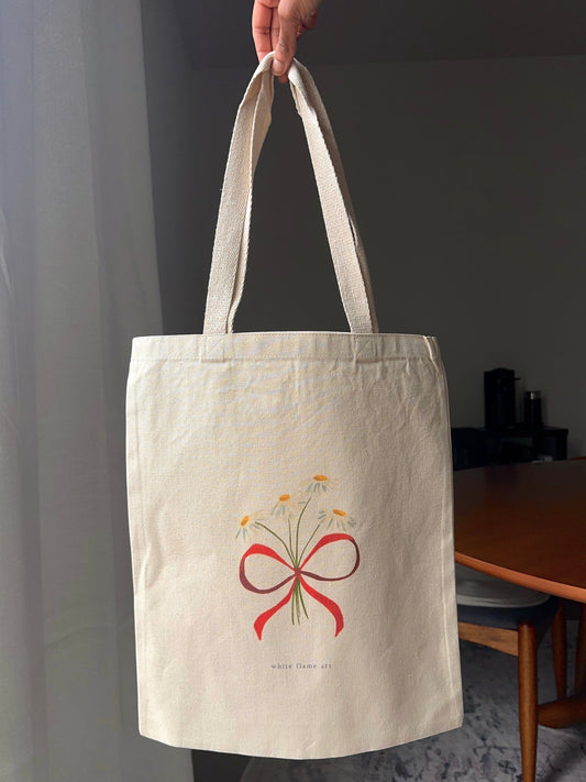 Beige tote bag with floral bow design held by a hand indoors.