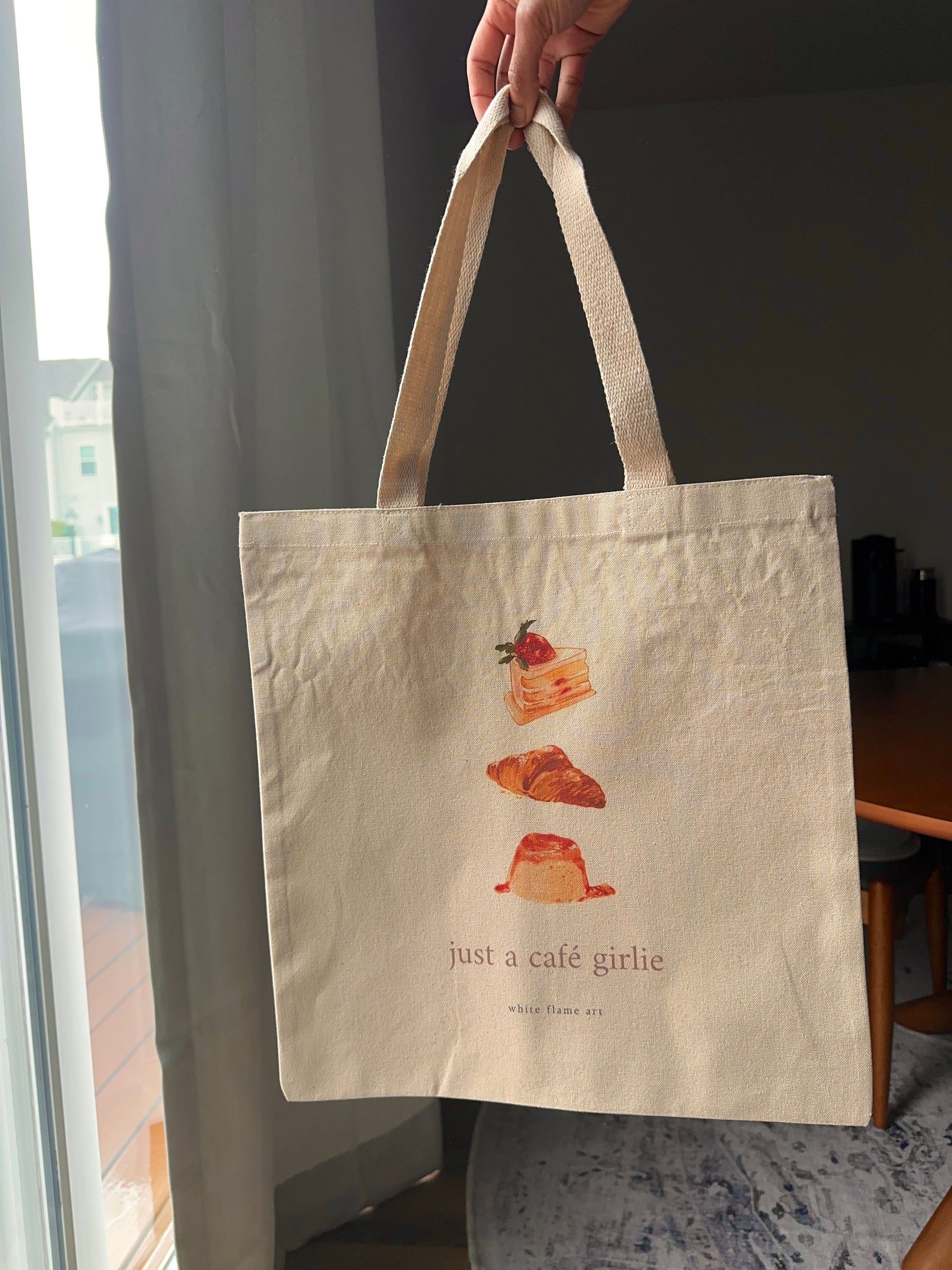 Tote bag with illustrations of pastries and text held by a hand indoors.