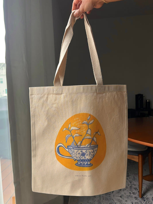 Beige tote bag with a teacup design held by a hand indoors.