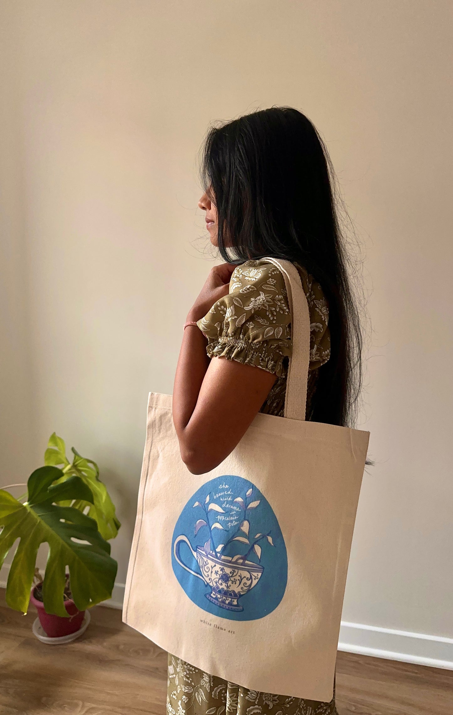 Woman holding a beige tote bag with a blue design, standing indoors.