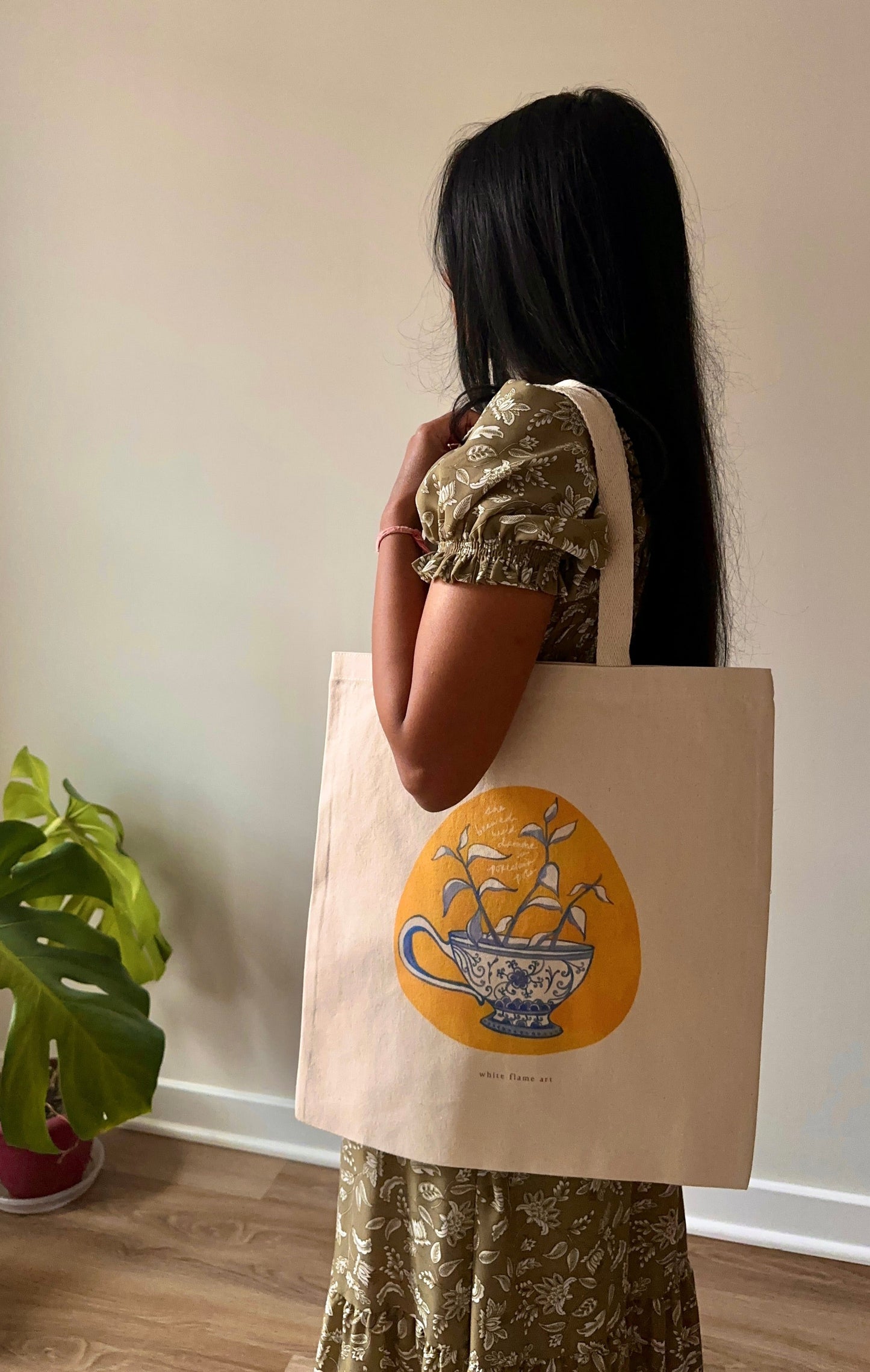 Person holding a tote bag with a design of a teacup and saucer, standing indoors next to a plant.